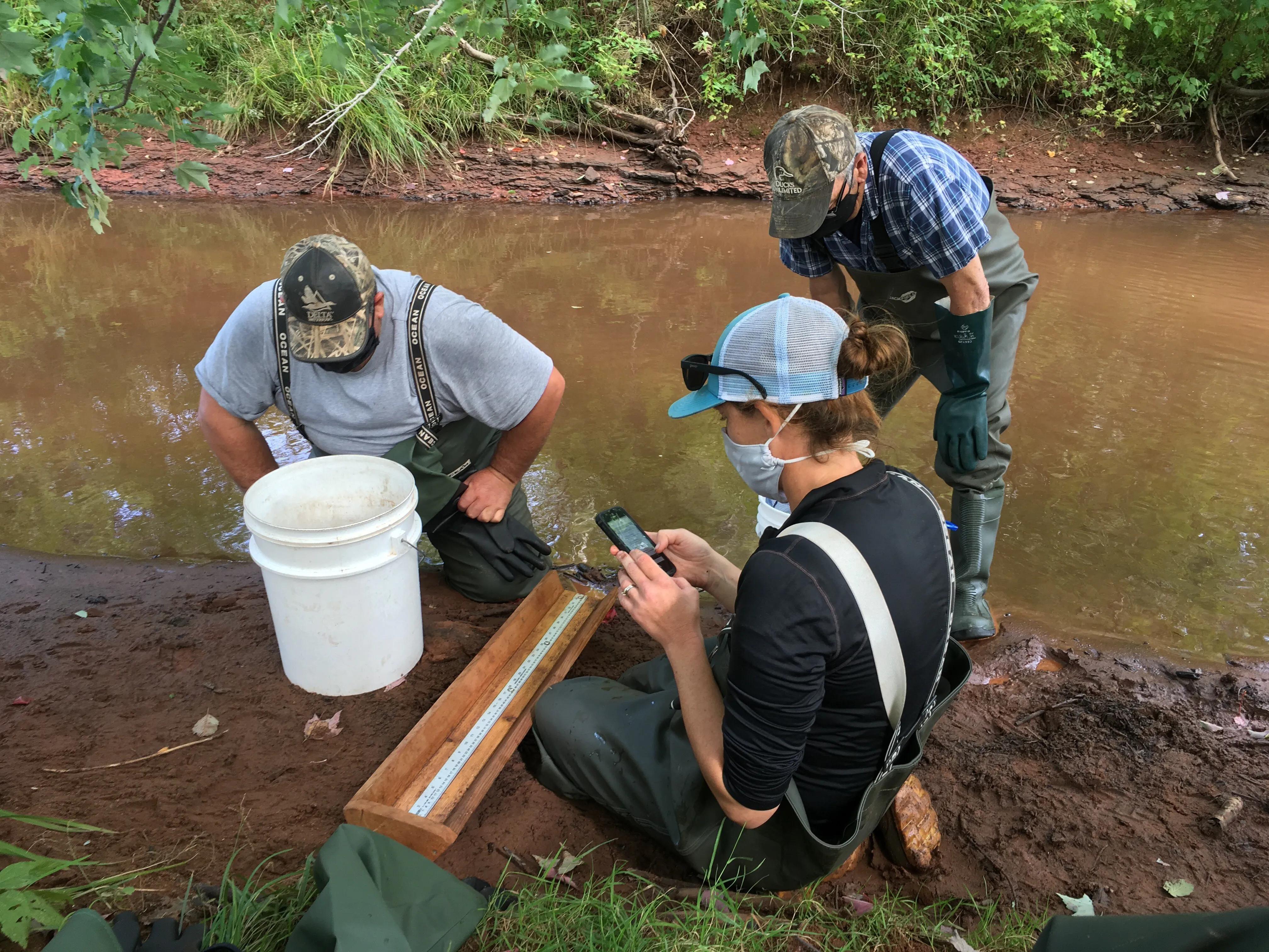 Three people standing by a riverstream analyzing water samples. Two are standing over a wooden instrument and one is looking at their phone. All three are wearing masks. CBM in Action; PEI Watershed Alliance