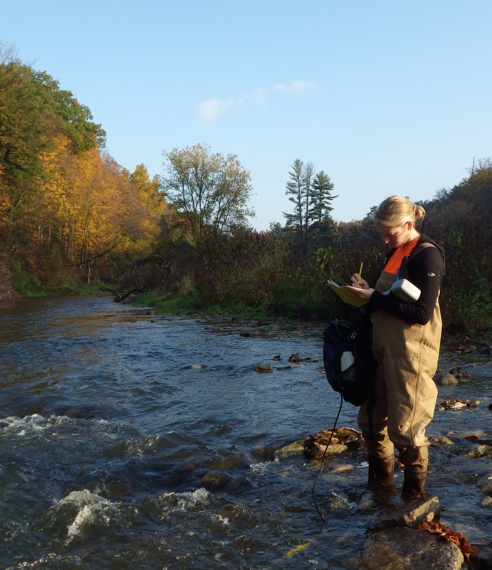 A woman in chest waders stands in the middle of a stream taking notes while holding a YSI meter that is in the water