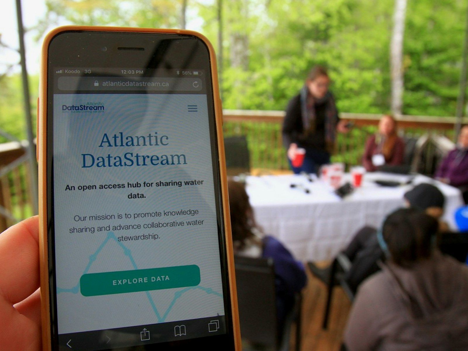  Iphone affichant la page d'accueil d'Atlantic DataStream avec des personnes sur une terrasse autour d'une table en arrière-plan