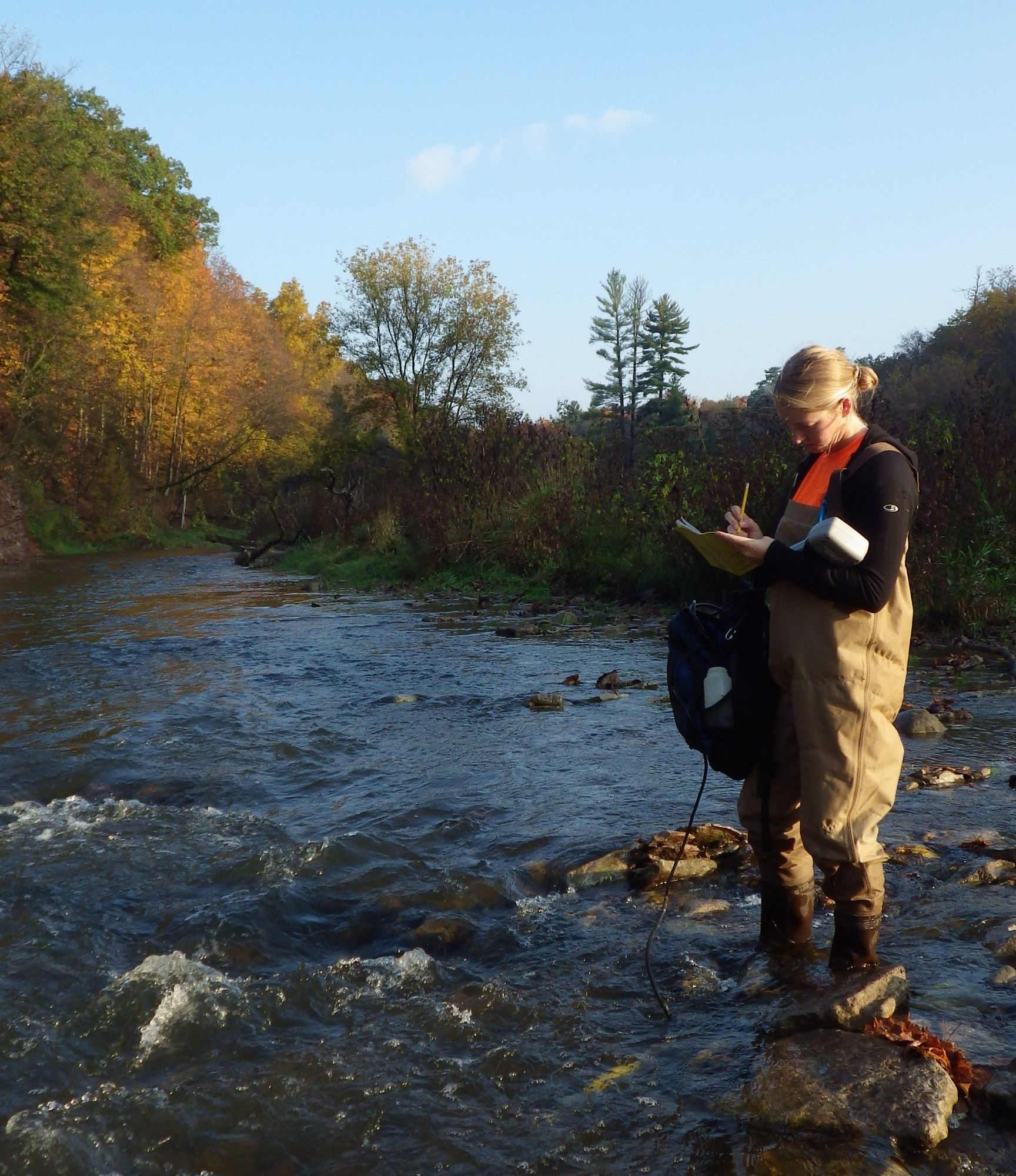 Une femme en bottes cuissardes se tient au milieu d’un ruisseau et prend des notes tout en tenant une sonde YSI qui est dans l’eau.