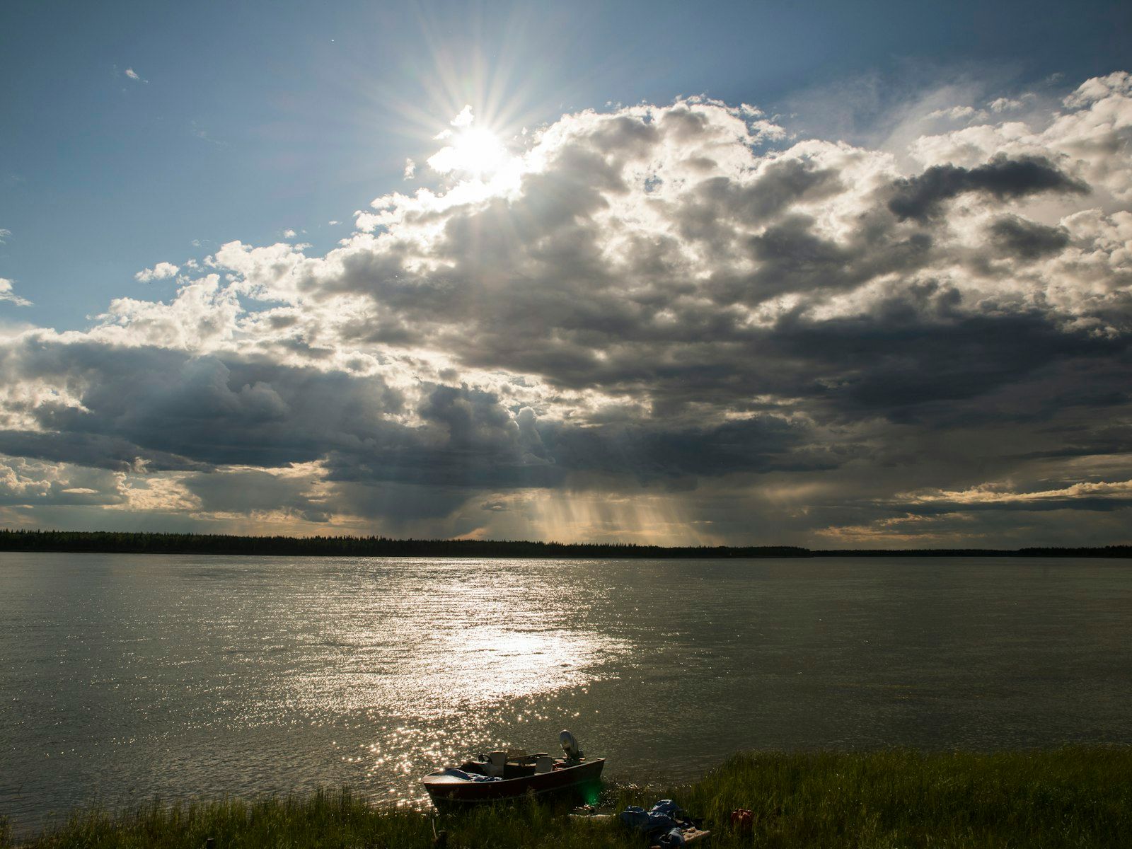Sun peaking out from behind clouds over a boat on the shoreline of a lake