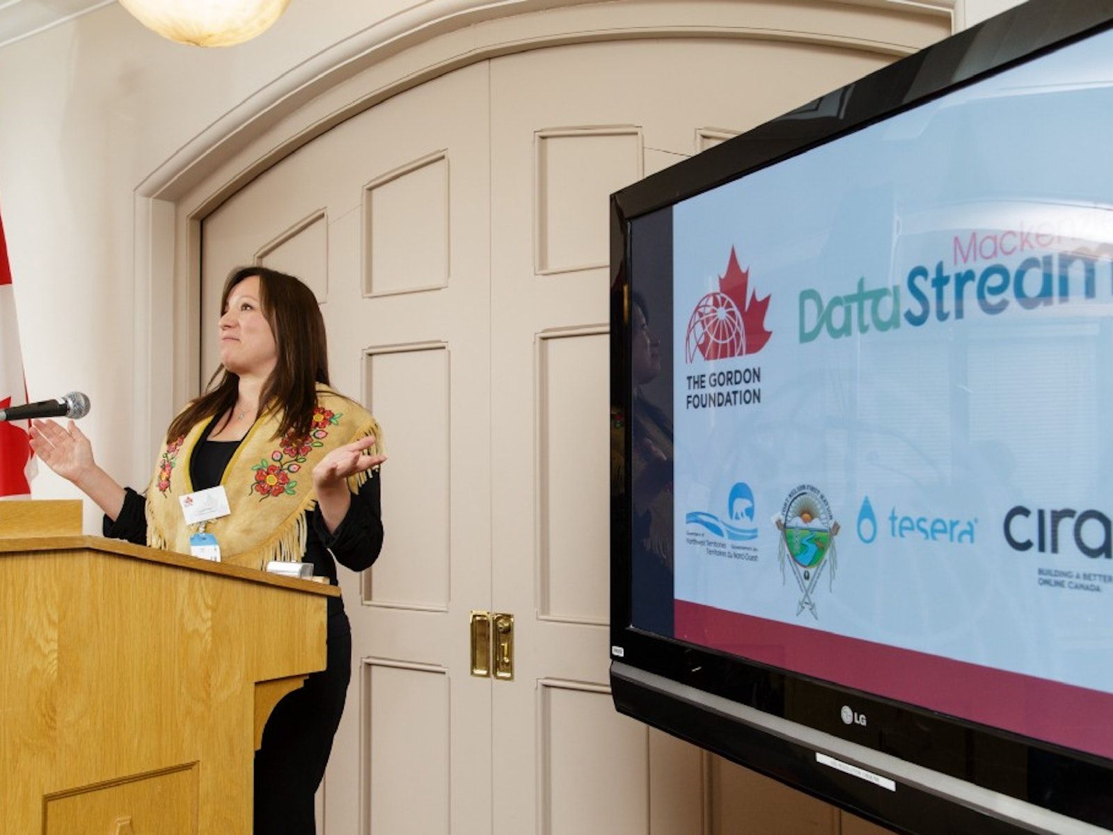 Lana Lowe at a podium next to a screen displaying logos for the gordon foundation, mackenzie datastream, the government of the northwest territories, Fortn Nelson First Nation, Tesera, and CIRA