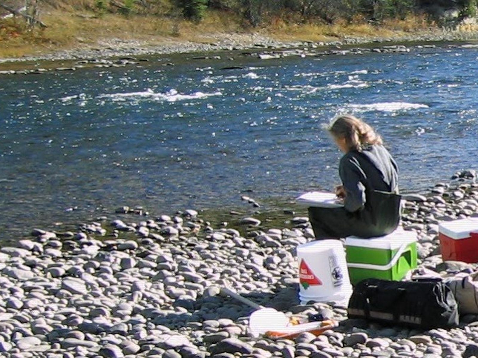 Woman sitting on a cooler next to a river recording water quality results onto a field sheet